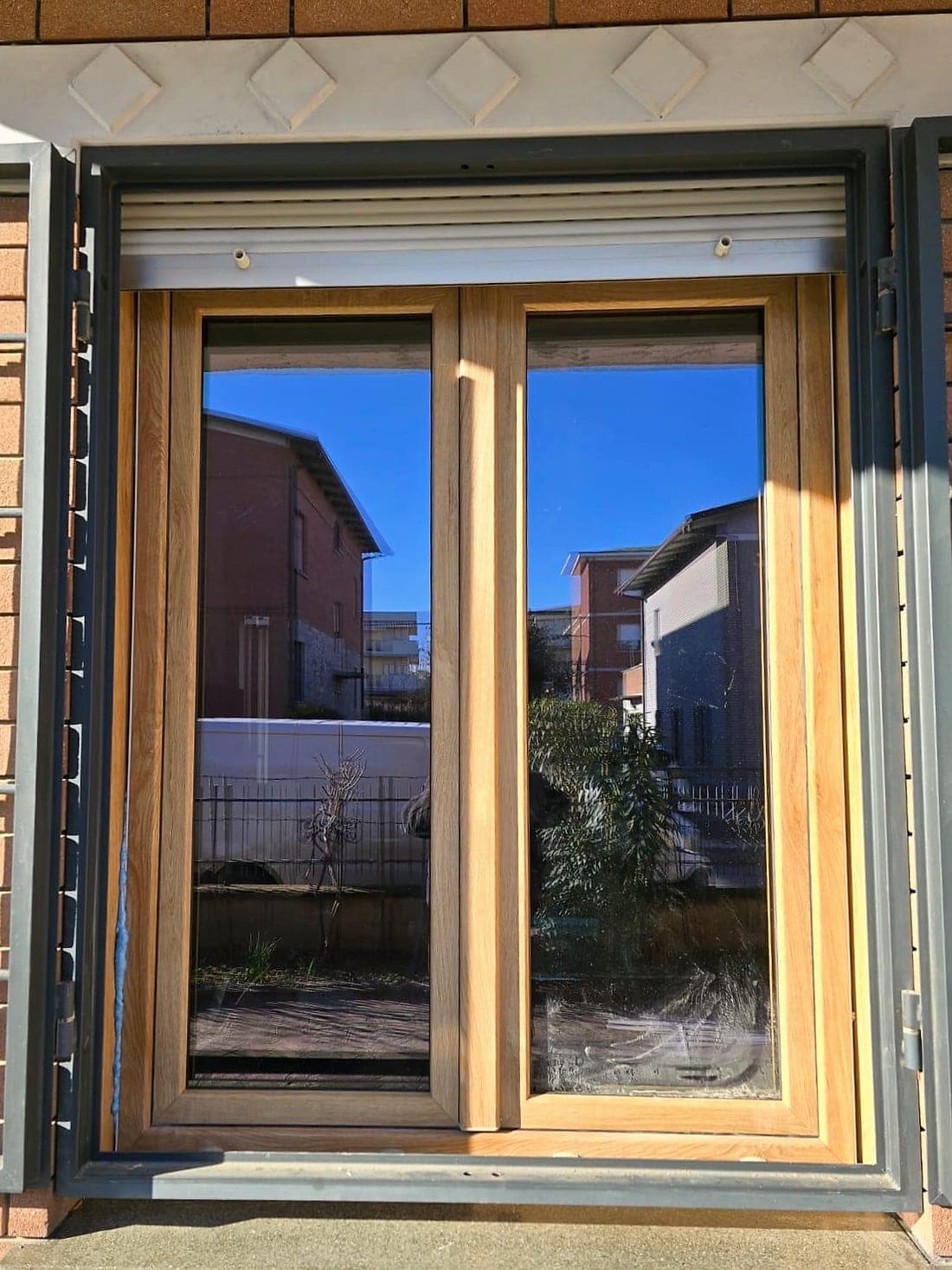 Finestra effetto rovere con tapparella bianca e grata di sicurezza antracite, vista dall'esterno